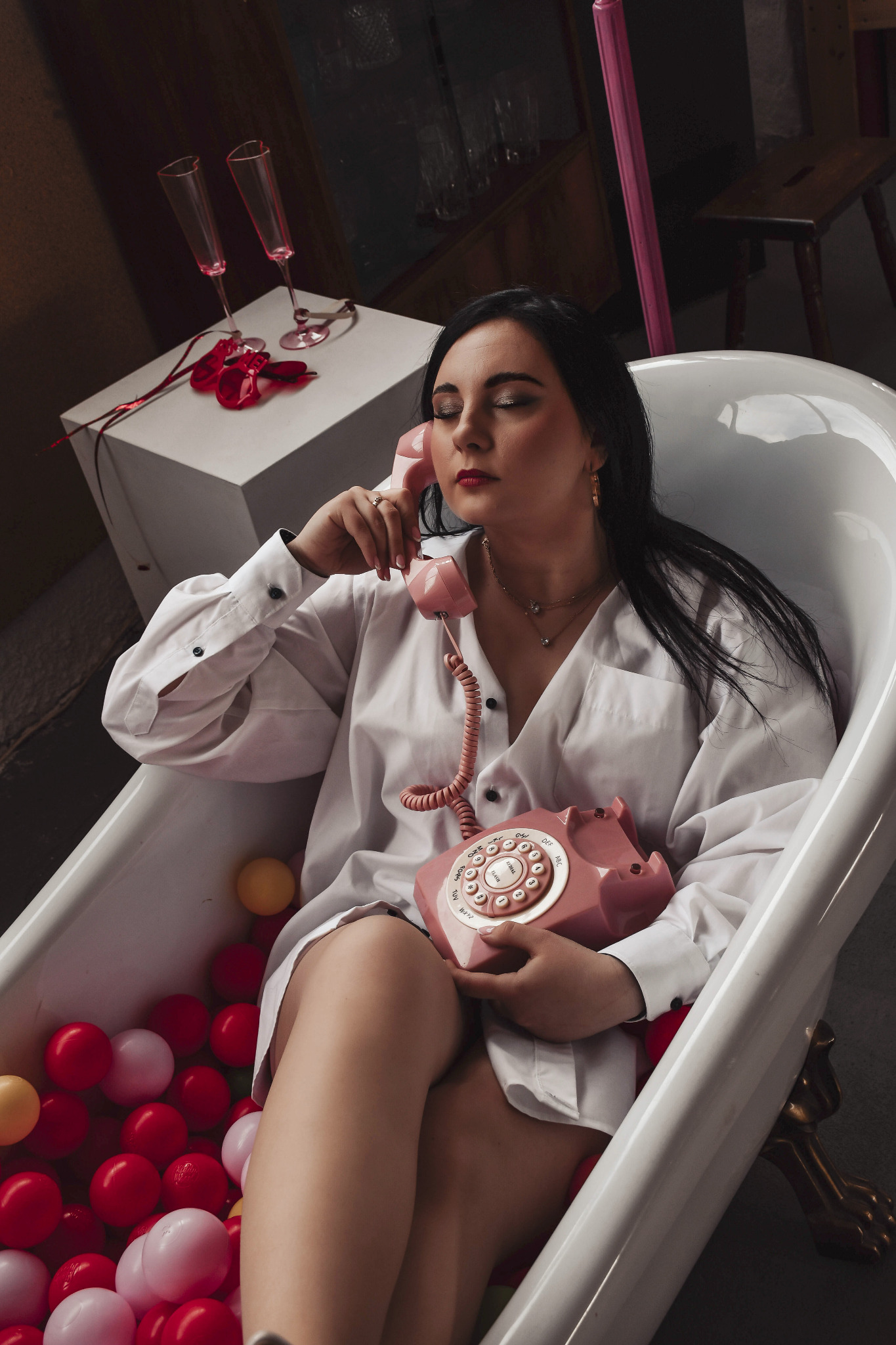 Kobieta w białej koszuli siedzi w wannie wypełnionej kolorowymi piłkami, trzymając różowy telefon retro. Na białym stoliku kieliszki i czerwone róże.
