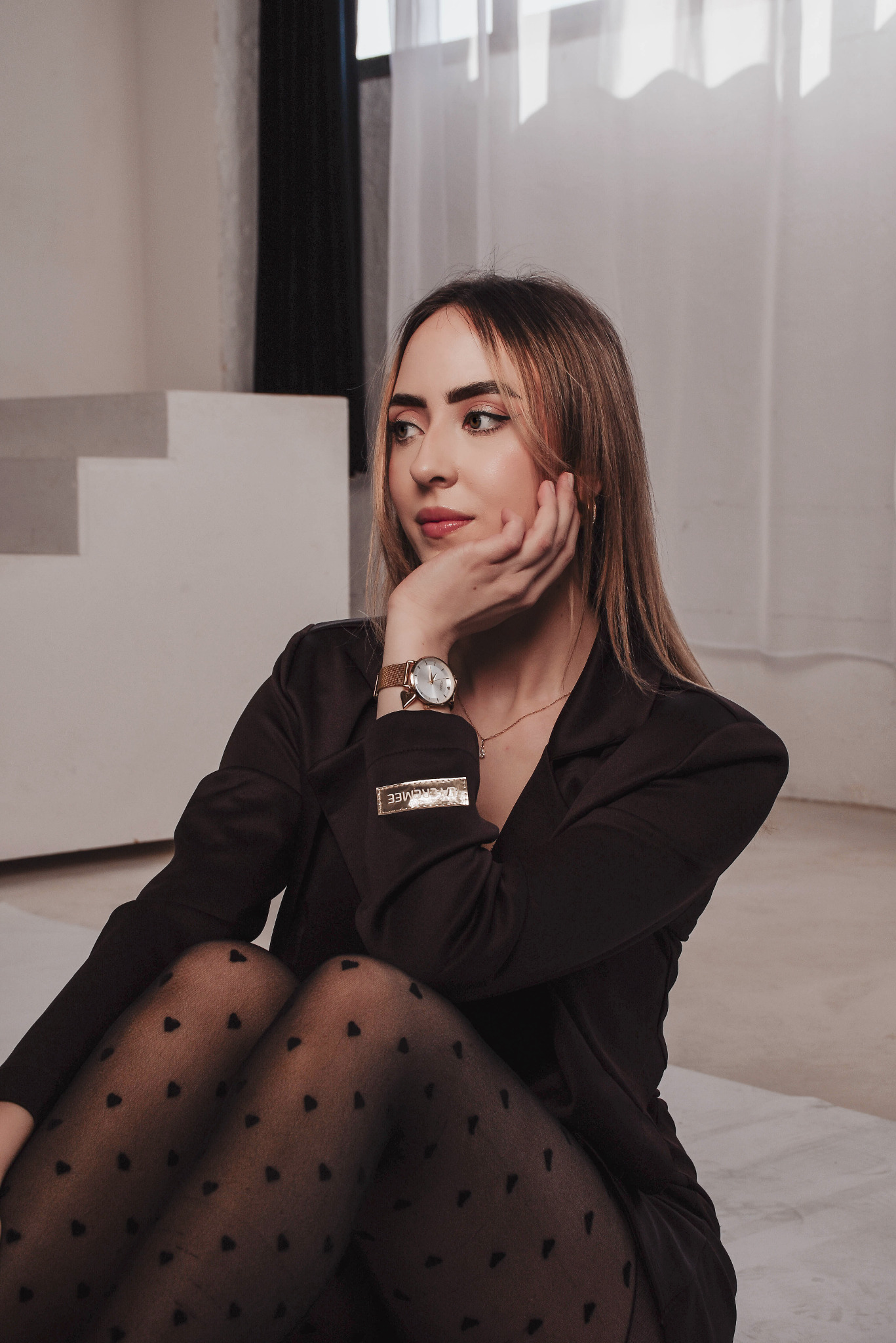 Portret kobiety w czarnym żakiecie i rajstopach w serduszka, z zegarkiem na ręku, siedzącej na podłodze w studio z minimalistycznym tłem.