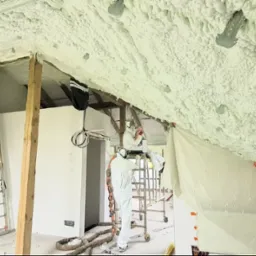 Prace izolacyjne poddasza pianką PUR: pracownicy w kombinezonach ochronnych aplikują pianę na skośny strop, widoczne rusztowania, folia ochronna i elementy konstrukcyjne dachu.