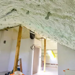 Poddasze z ociepleniem pianką PUR, widoczne metalowe elementy mocujące, drewniany słup i okno dachowe. Ściany w trakcie wykończenia, widoczne instalacje.