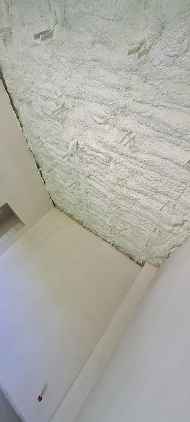 Poddasze po aplikacji białej pianki izolacyjnej, widoczne elementy mocujące. Układ warstw izolacji i surowy stan ścian w jasnym wnętrzu.