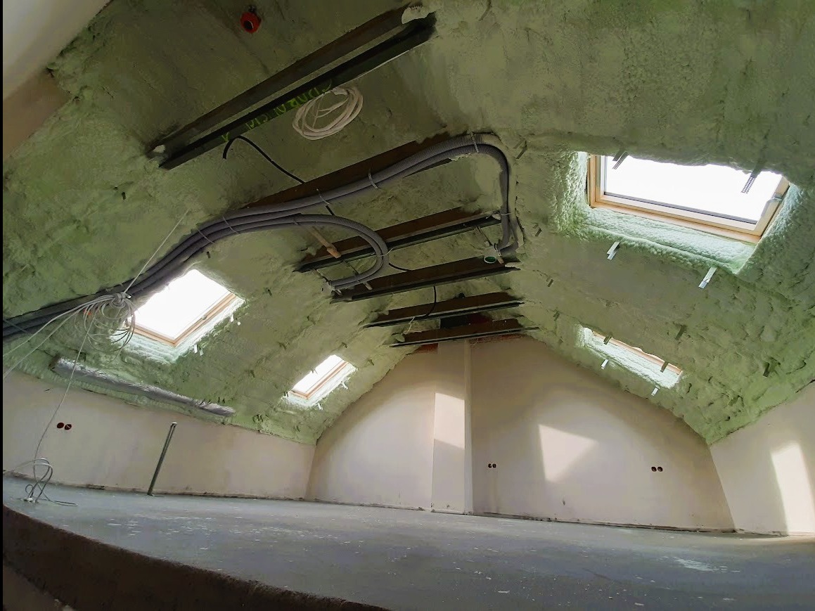 Poddasze po ociepleniu pianką PUR z widocznymi oknami dachowymi i przewodami instalacyjnymi. Ściany częściowo otynkowane, surowa podłoga.