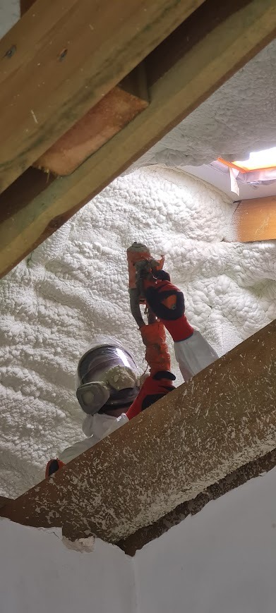 Pracownik w kombinezonie ochronnym aplikuje piankę PUR na poddasze. Widoczna struktura pianki i drewniane elementy konstrukcyjne dachu.