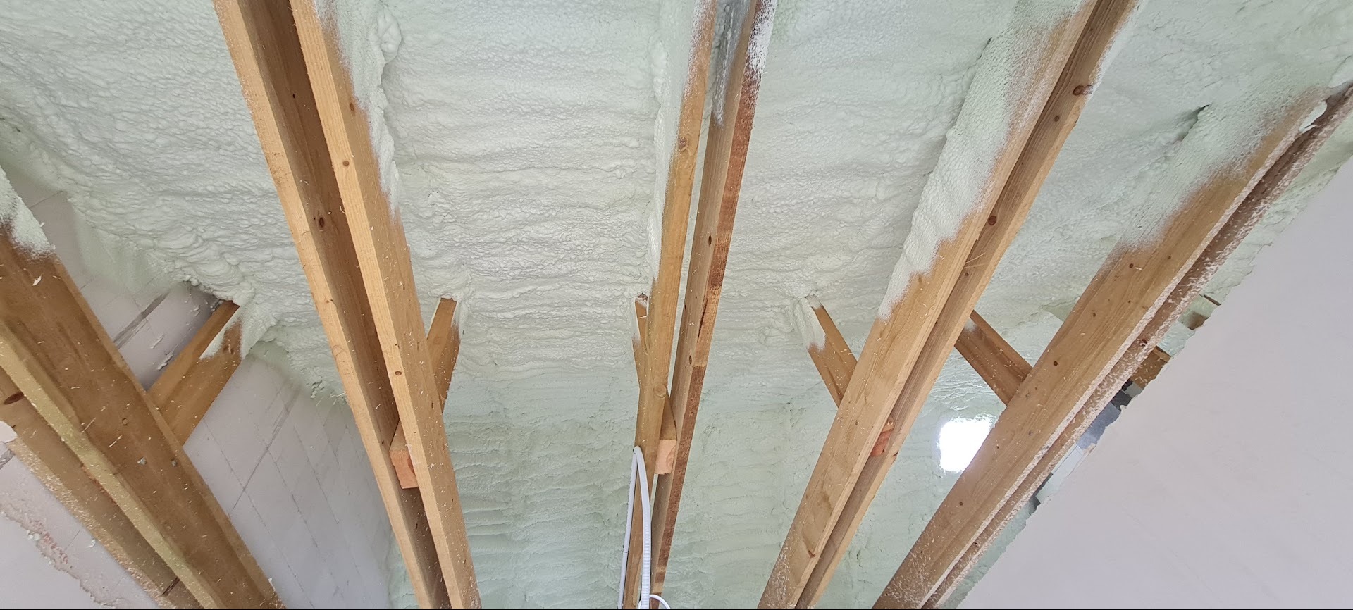 Poddasze z drewnianą konstrukcją pokryte warstwą jasnej pianki izolacyjnej, widok z dołu, nierównomierna powierzchnia piany.