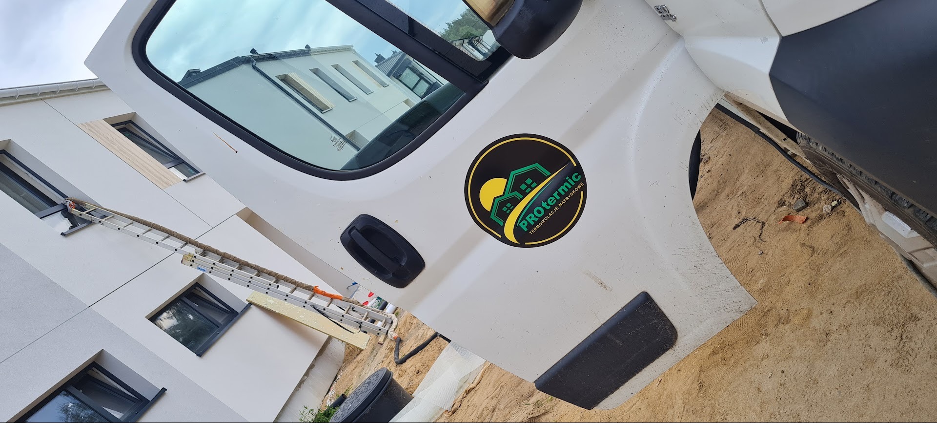 Biały samochód z naklejką firmy Protermic na tle ocieplanego budynku z drabiną. Widoczna fasada i okna w trakcie prac.