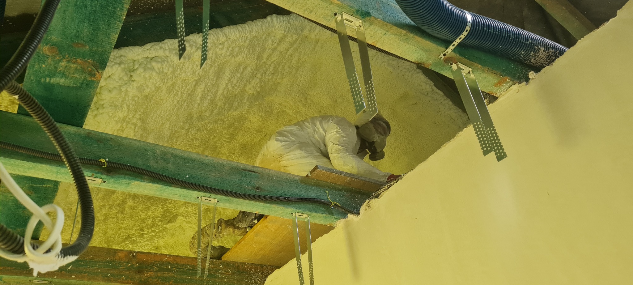 Pracownik w kombinezonie i masce aplikuje pianę PUR na poddasze, widoczne drewniane elementy konstrukcji i instalacja wentylacyjna.