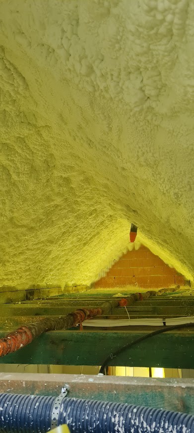 Poddasze ocieplone pianką PUR, widoczne krokwie i fragment ściany z cegły. Aplikacja pianki o nieregularnej strukturze, rury instalacyjne w dolnej części kadru.