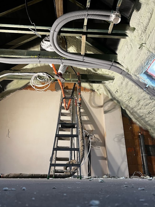 Prace izolacyjne na poddaszu: aplikacja pianki PUR na skośnych ścianach. Widoczne przewody wentylacyjne i drabina. Detal: białe fragmenty pianki na podłodze.