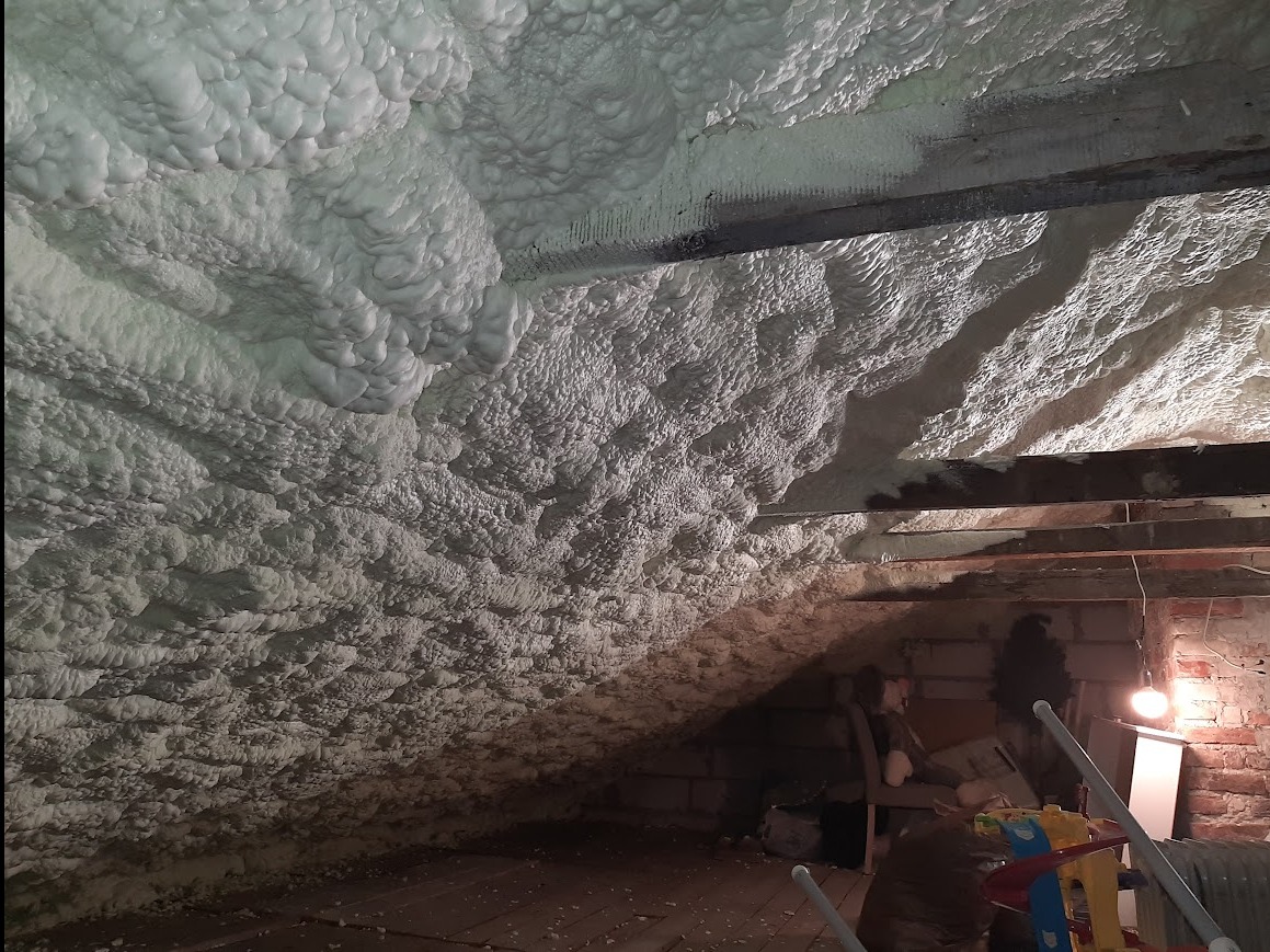 Poddasze po izolacji pianką PUR, widoczne drewniane belki i nierównomierna struktura piany. Oświetlenie żarówką podkreśla fakturę.