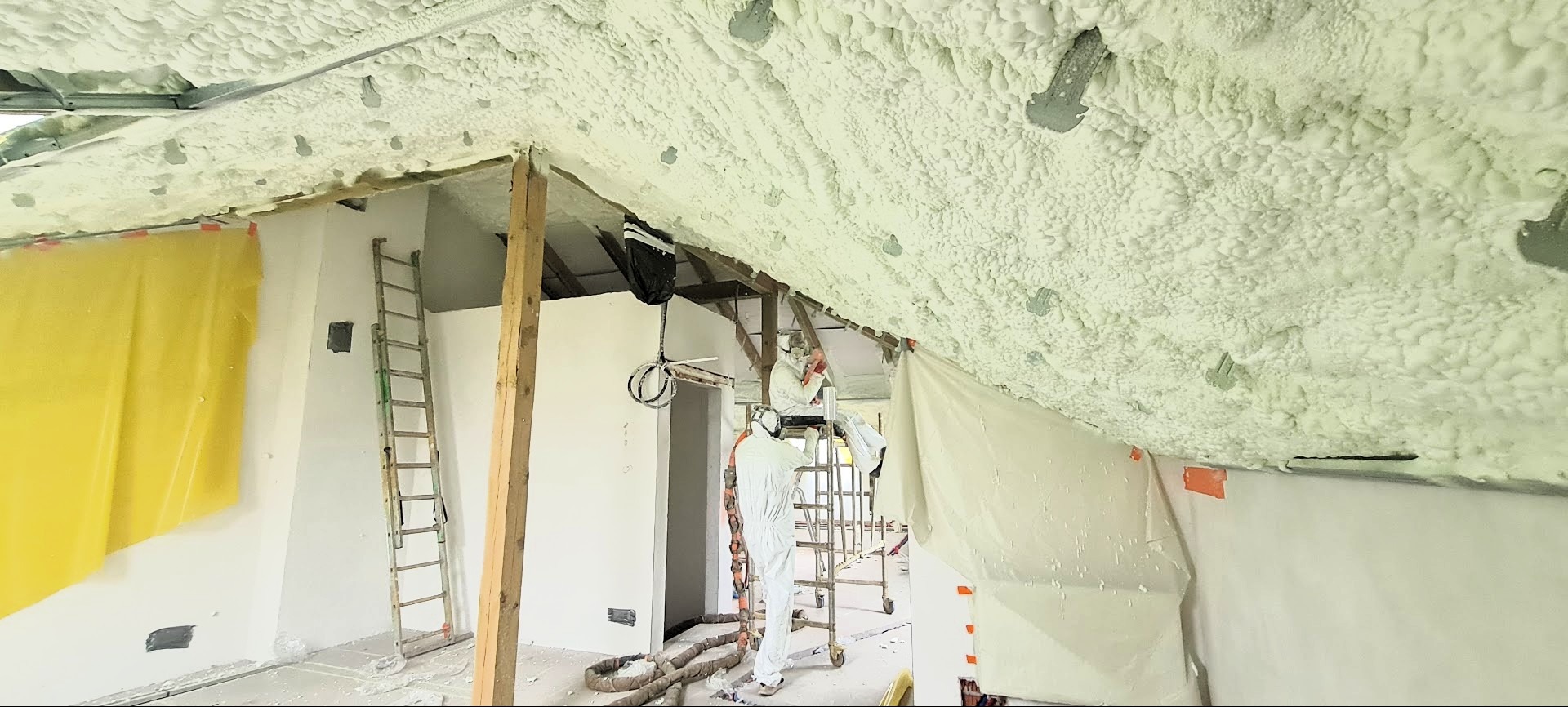 Prace izolacyjne poddasza pianką PUR: pracownicy w kombinezonach ochronnych aplikują pianę na skośny strop, widoczne rusztowania, folia ochronna i elementy konstrukcyjne dachu.