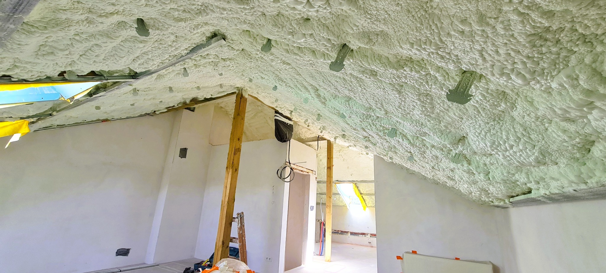 Poddasze z ociepleniem pianką PUR, widoczne metalowe elementy mocujące, drewniany słup i okno dachowe. Ściany w trakcie wykończenia, widoczne instalacje.