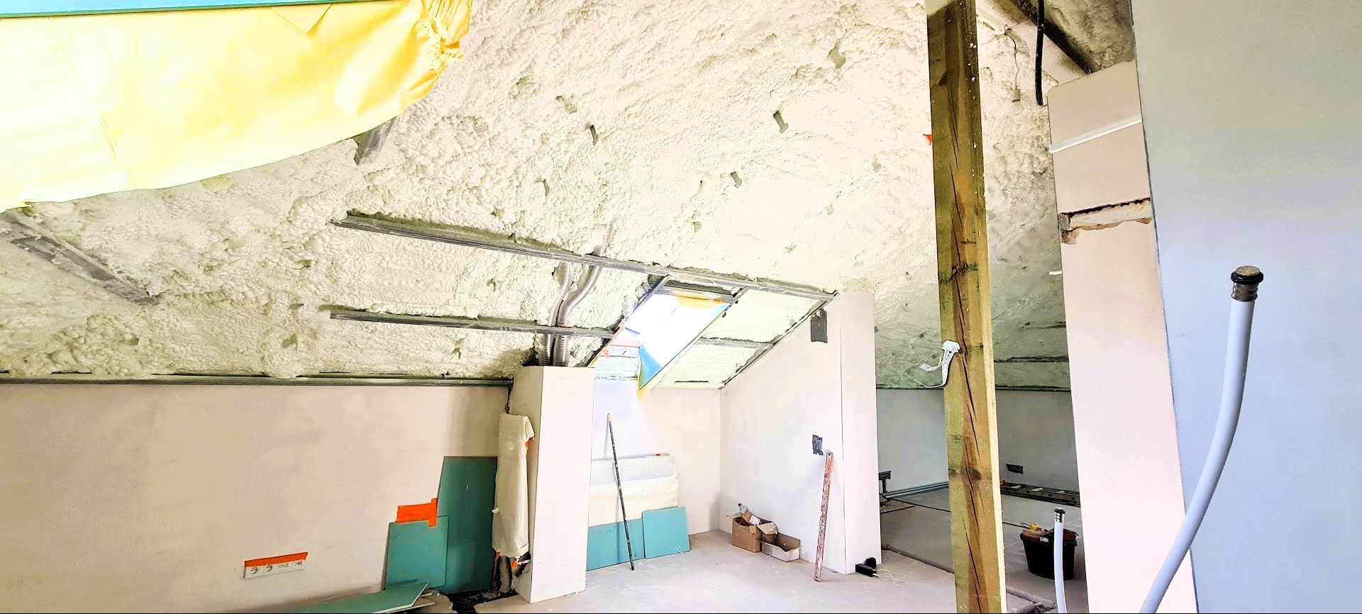 Poddasze po natrysku pianki PUR, widoczne metalowe profile, instalacje i fragmenty ścian. Na podłodze płyty gipsowe i narzędzia. Oświetlenie naturalne, wnętrze w trakcie prac.
