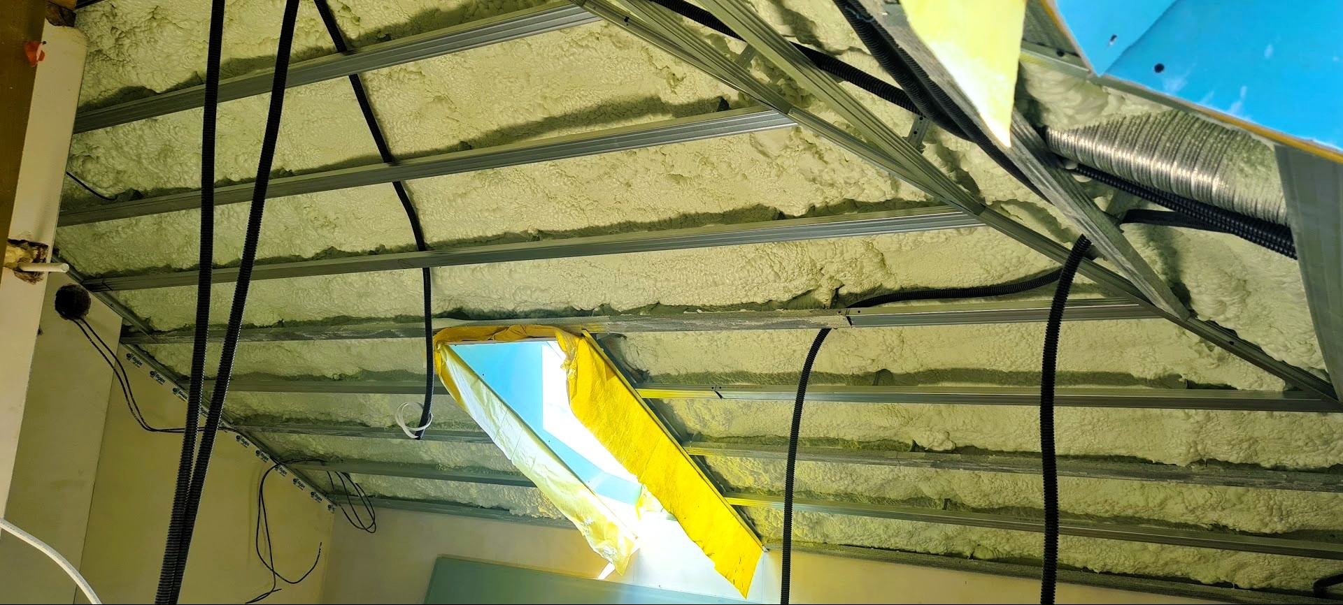 Poddasze z widoczną pianką izolacyjną między metalowymi profilami, czarne rury instalacyjne. Okno dachowe zabezpieczone folią.