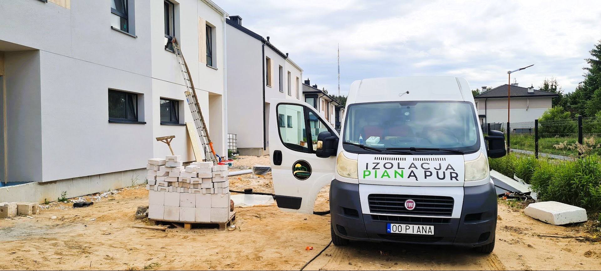 Prace izolacyjne na budynku: biały bus z logo firmy, pracownik na drabinie przy oknie, paleta z bloczkami. Widok na elewację w trakcie ocieplania.