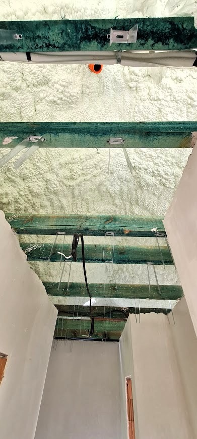 Poddasze po ociepleniu pianką PUR, widoczne zielone belki konstrukcyjne z metalowymi uchwytami i instalacja elektryczna. Pianka o nierównej strukturze.