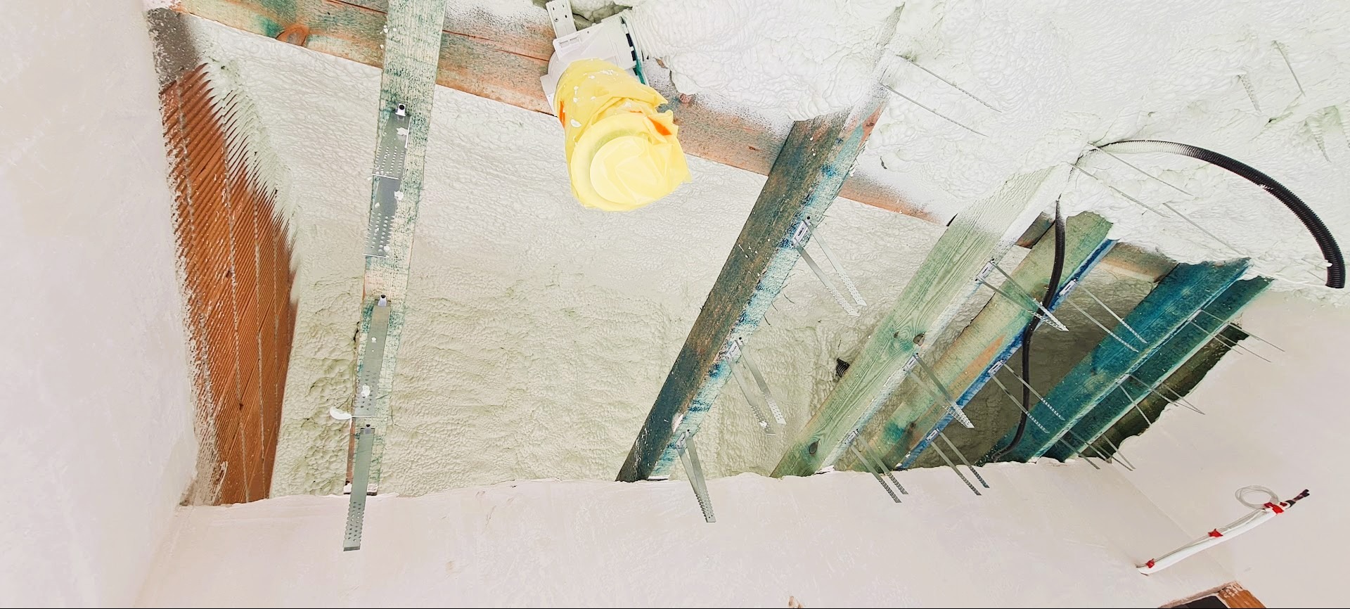 Poddasze ocieplone pianką PUR, widoczne drewniane elementy konstrukcyjne i żółty kask. Biała piana pokrywa skośne powierzchnie.