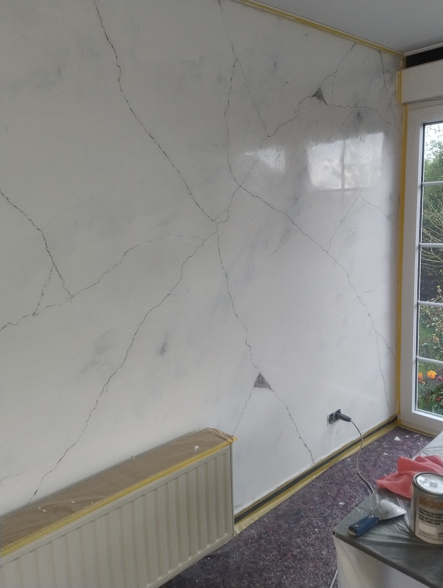 Ściana w pokoju po dekoracyjnym malowaniu imitującym marmurkowe żyłki. Grzejnik i podłoga zabezpieczone folią i taśmą. Widoczne narzędzia malarskie.