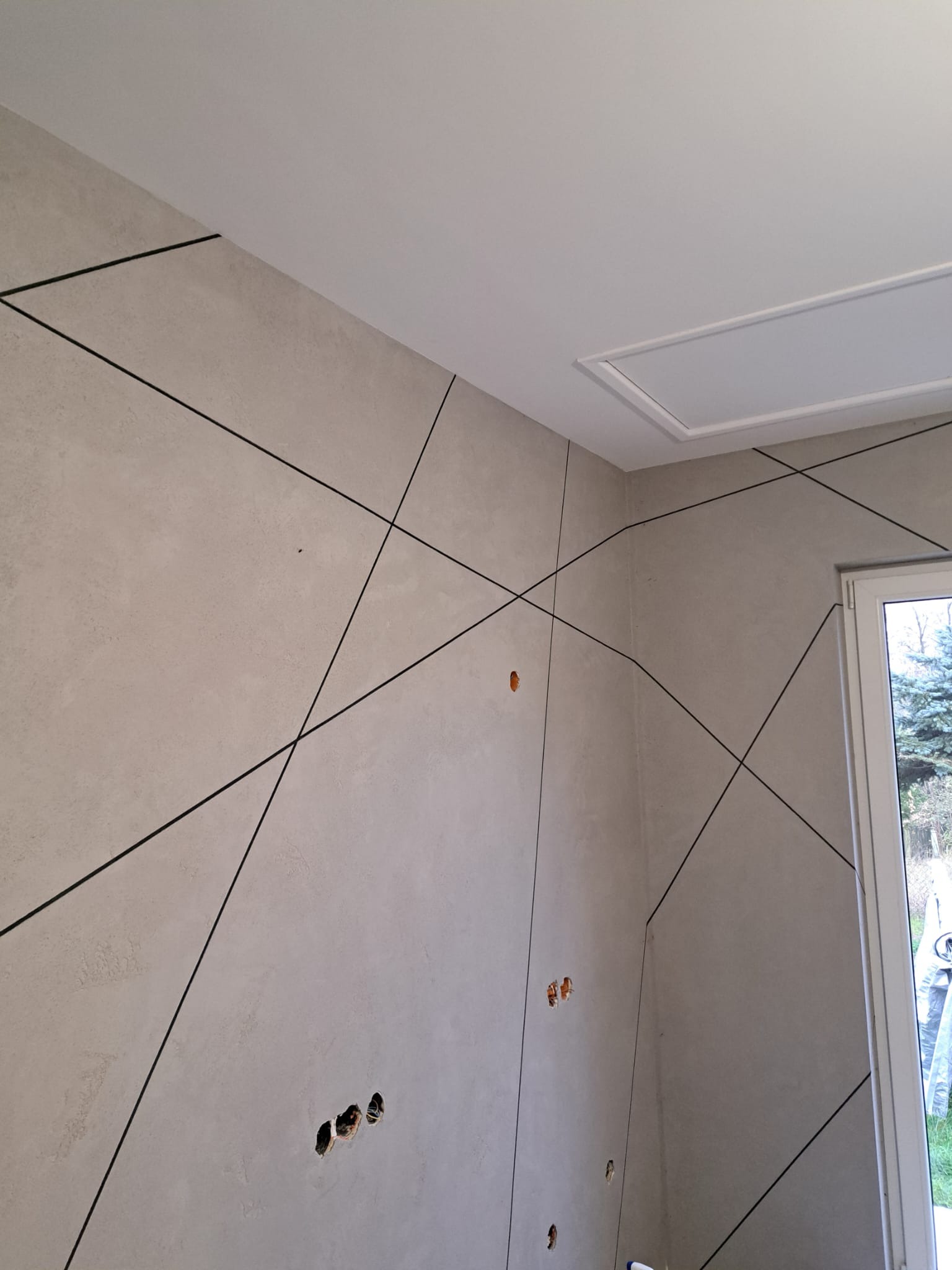 Ściana z beżową strukturą i czarnymi liniami tworzącymi geometryczny wzór. Widoczne otwory na gniazdka i włączniki, sufit z klapą rewizyjną i fragment okna.