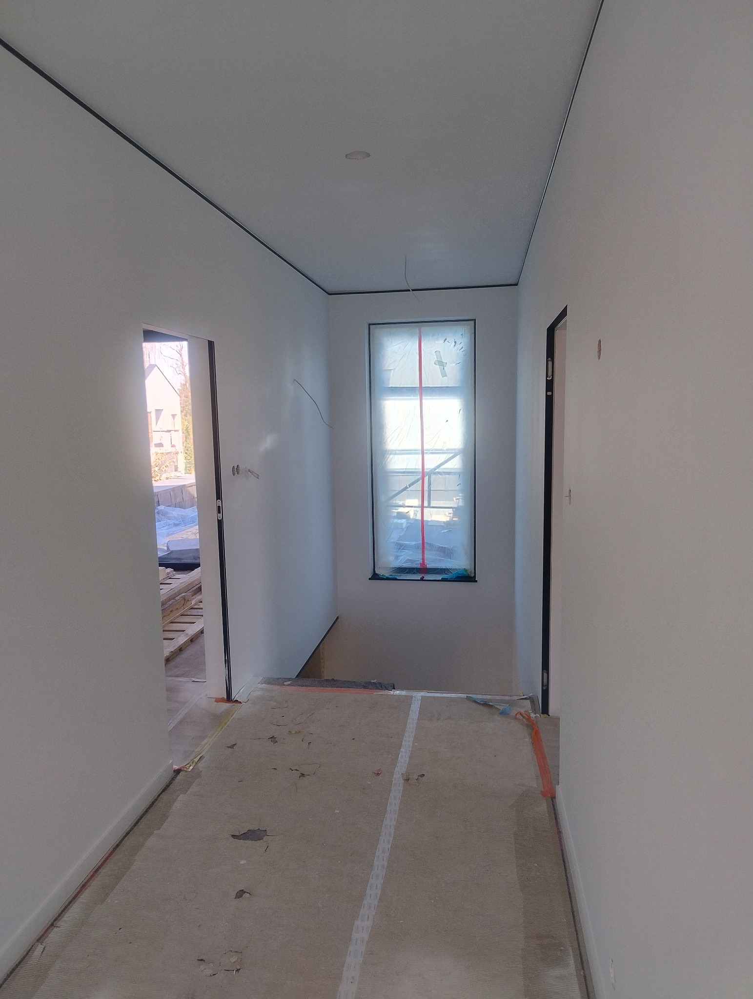 Korytarz w trakcie malowania na biało, okno zabezpieczone folią. Widoczne listwy sufitowe i przygotowane podłoże. Prace wykończeniowe w nowym domu.