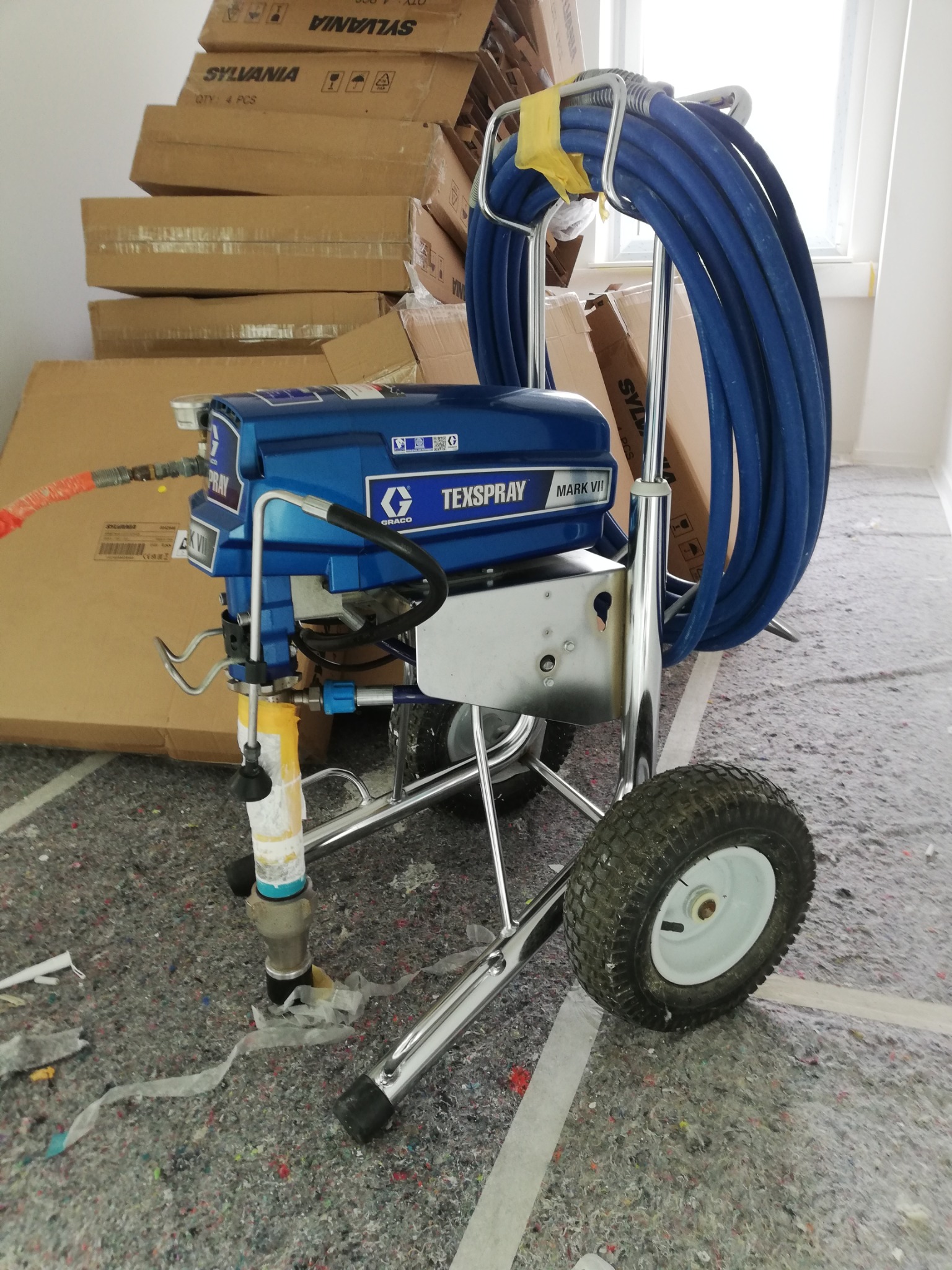 Agregat malarski Graco Texspray Mark VII na kółkach, z niebieskim wężem, stoi na zabezpieczonej folią podłodze w pomieszczeniu z kartonami w tle. Przygotowanie do malowania.