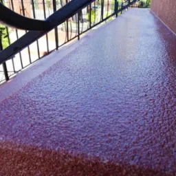 Czerwona, teksturowana powierzchnia balkonu z metalową balustradą w tle, wykonana w technologii imitującej kamień.