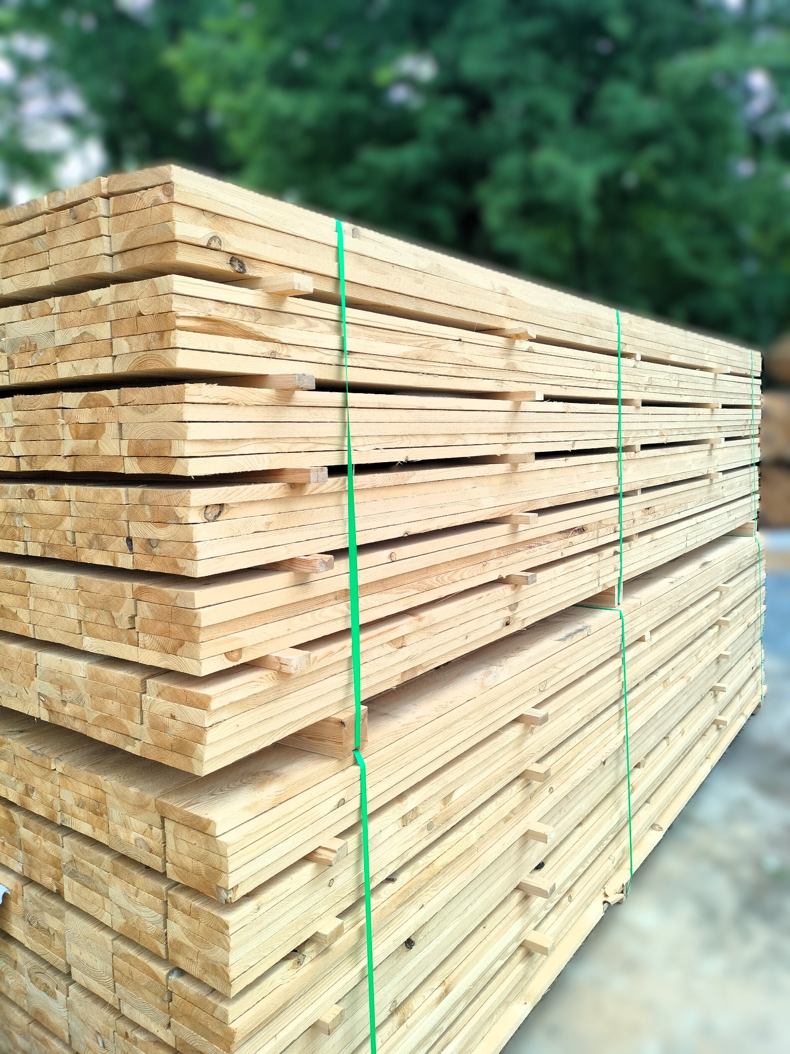 Stos tarcicy budowlanej z zielonymi taśmami, widoczne słoje drewna i drobne sęki. Tło rozmyte, zielone drzewa. Lekki kąt z dołu podkreśla objętość materiału.