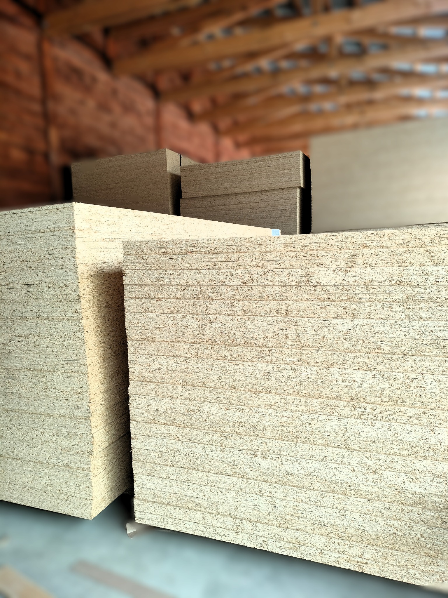 Stosy płyt OSB w magazynie, widoczne krawędzie i faktura materiału. W tle konstrukcja drewniana dachu. Ujęcie z bliska podkreśla strukturę płyt.