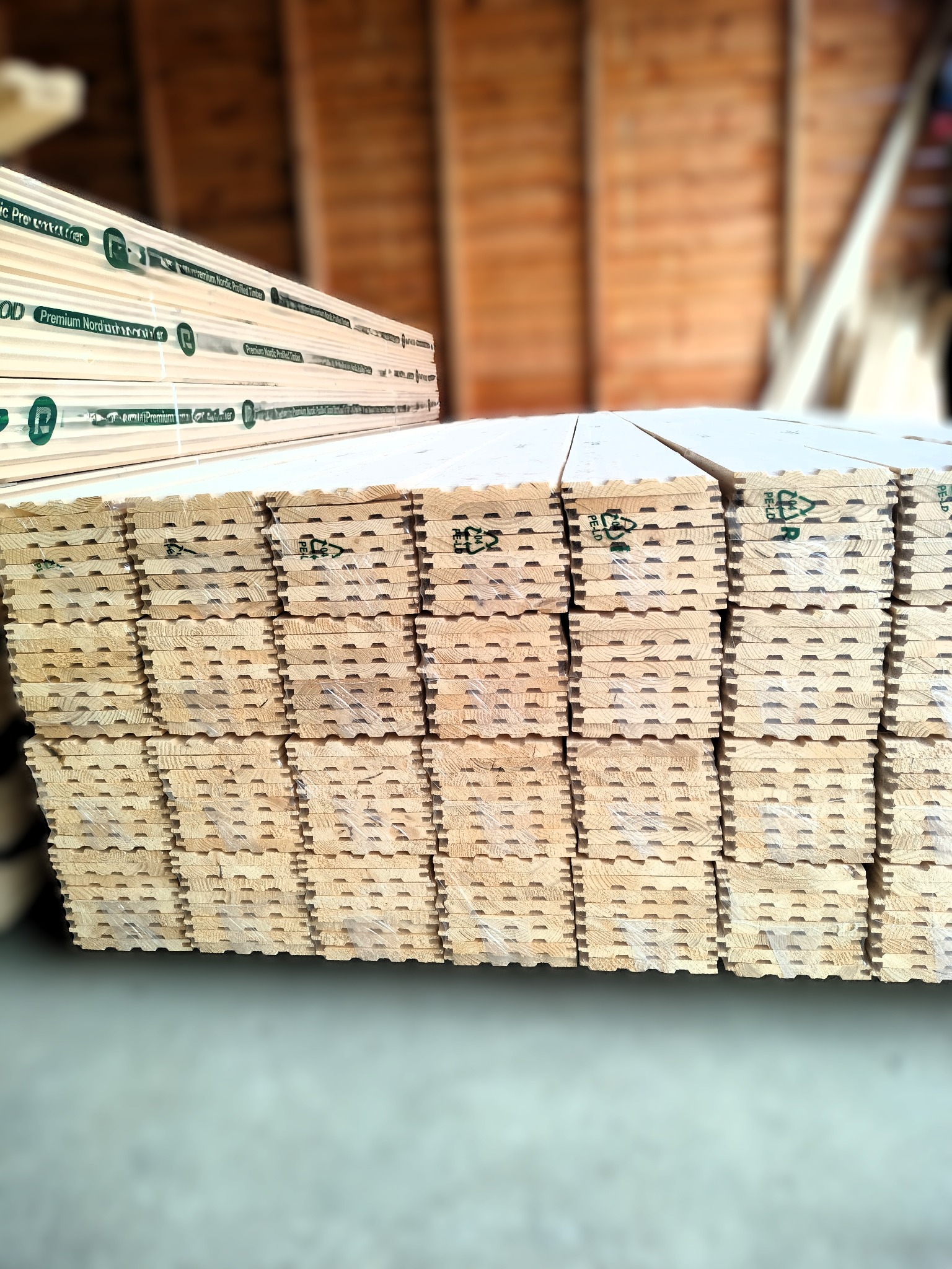 Stos tarcicy profilowanej Premium Nordic Timber, zabezpieczonej folią. Widoczne oznaczenia PE-LD. W tle magazyn drewna z deskami na regałach.