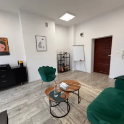 Jasne wnętrze gabinetu z zielonymi fotelami i drewnianymi stolikami. Na ścianach wiszą plakaty, a w rogu stoi tablica suchościeralna. Przytulna przestrzeń do rozmów.