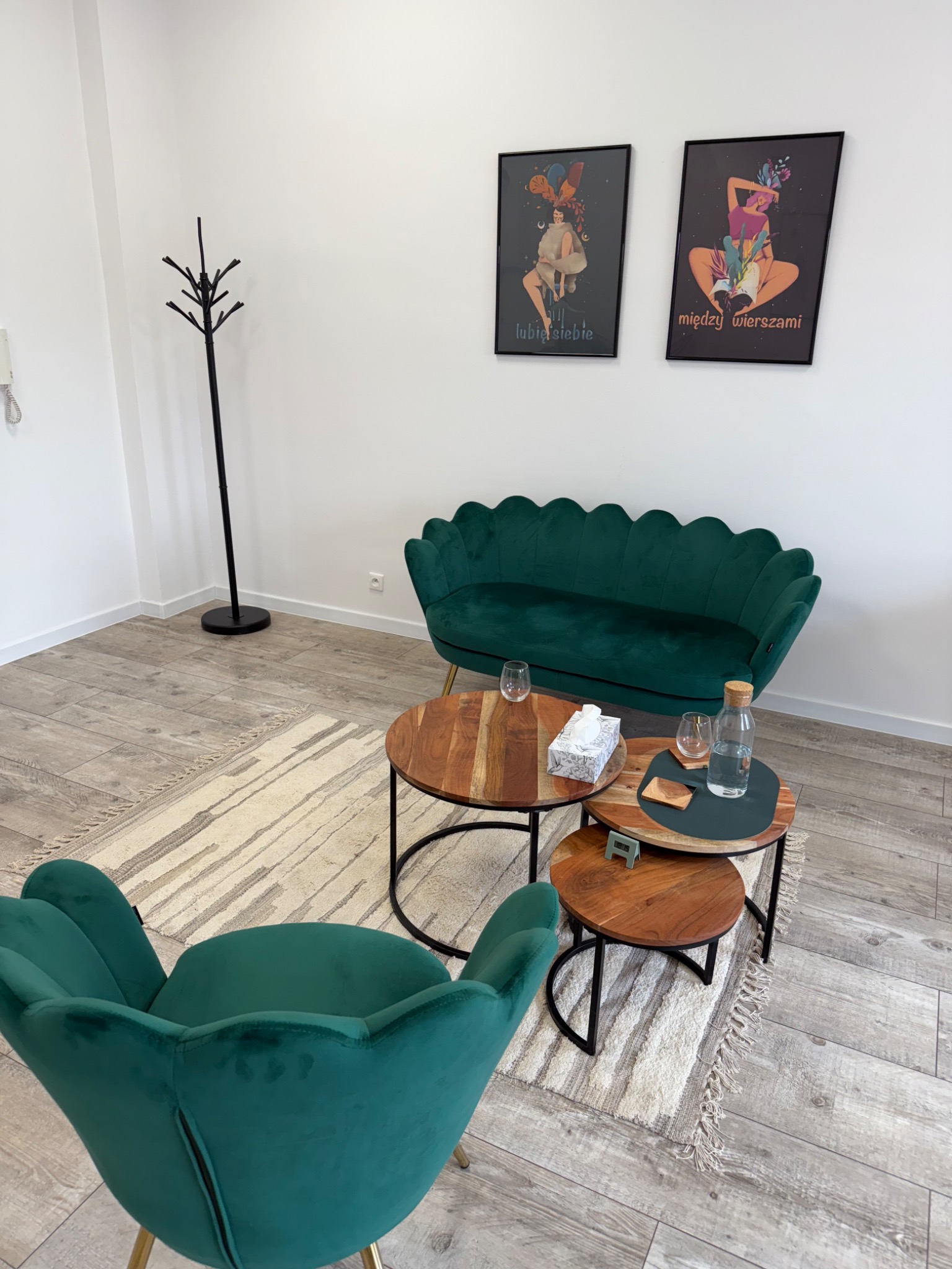 Przytulny gabinet: szmaragdowa sofa i fotel w kształcie kwiatu, drewniane stoliki kawowe, dywan w paski, plakaty na ścianie. Jasne, minimalistyczne wnętrze.