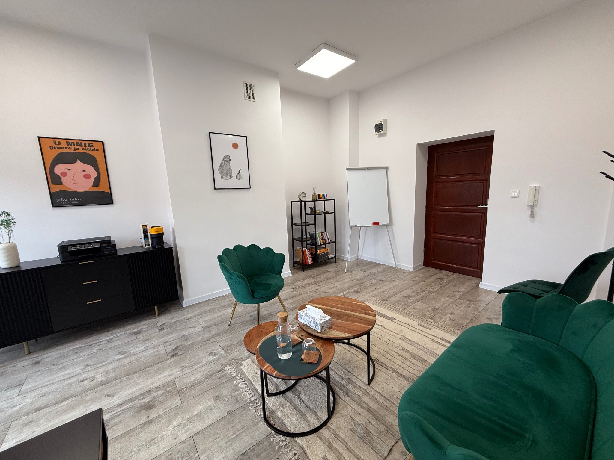 Jasne wnętrze gabinetu z zielonymi fotelami i drewnianymi stolikami. Na ścianach wiszą plakaty, a w rogu stoi tablica suchościeralna. Przytulna przestrzeń do rozmów.