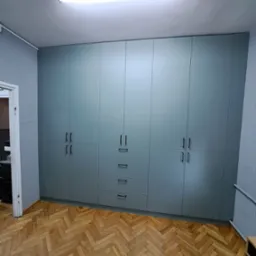 Szara szafa wnękowa z czarnymi uchwytami, widok z dołu, parkiet w jodełkę, drzwi po lewej stronie, oświetlenie sufitowe.