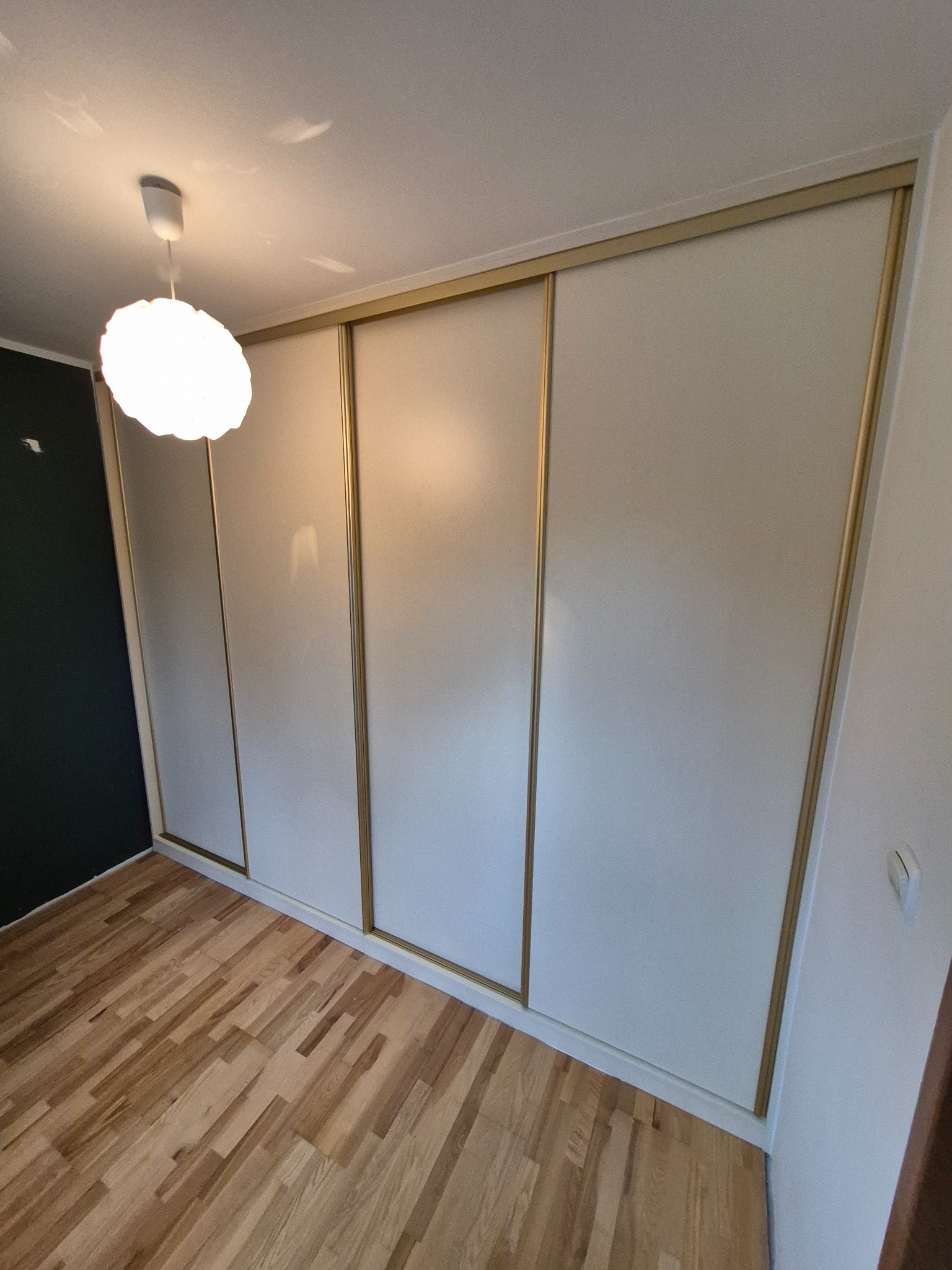 Biała szafa wnękowa z przesuwnymi drzwiami i złotymi obramowaniami, widoczna w pokoju z drewnianą podłogą i lampą.