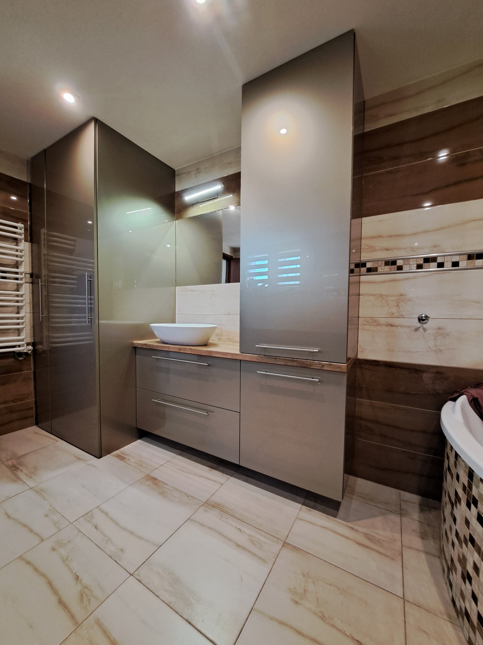 Nowoczesna, szara zabudowa łazienkowa z umywalką nablatową i wysokimi szafkami, dopasowana do płytek w odcieniach brązu i beżu. Całość dopełnia lustro.