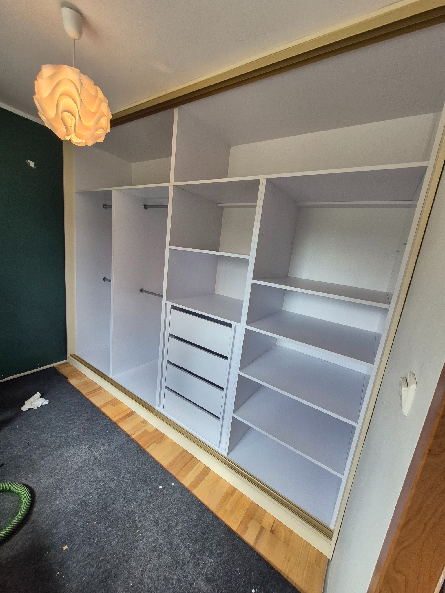 Biała szafa wnękowa z szufladami i półkami, wkomponowana w skos dachu. Widoczne metalowe drążki na ubrania i oświetlenie sufitowe.