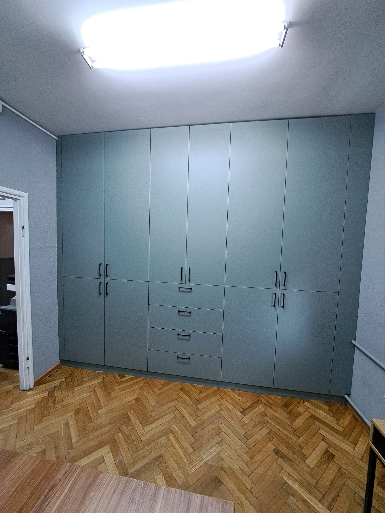 Szara szafa wnękowa z czarnymi uchwytami, widok z dołu, parkiet w jodełkę, drzwi po lewej stronie, oświetlenie sufitowe.