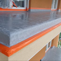 Firma Handlowo-Usługowa DOM Rojek Józef - Szara, błyszcząca posadzka na balkonie, zabezpieczona pomarańczową folią malarską, widok z góry. Na drugim planie elewacja budynku w odcieniach beżu i brązu.