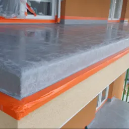 Szara, błyszcząca posadzka na balkonie, zabezpieczona pomarańczową folią malarską, widok z góry. Na drugim planie elewacja budynku w odcieniach beżu i brązu.