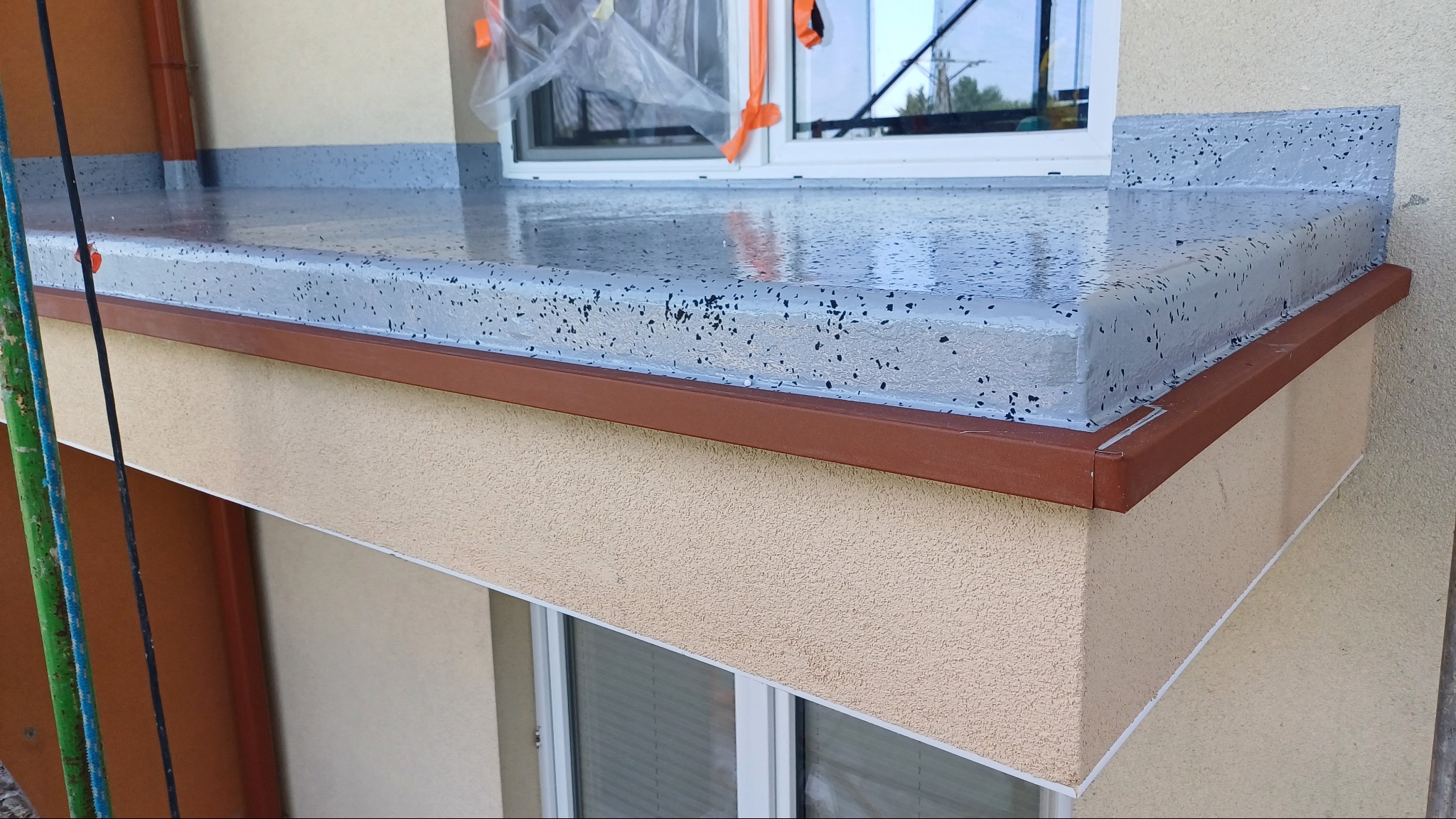 Balkon z szarą, połyskującą powierzchnią żywiczną z czarnymi drobinkami, wykończony brązową listwą. Widoczne okno i elewacja budynku w neutralnym odcieniu.