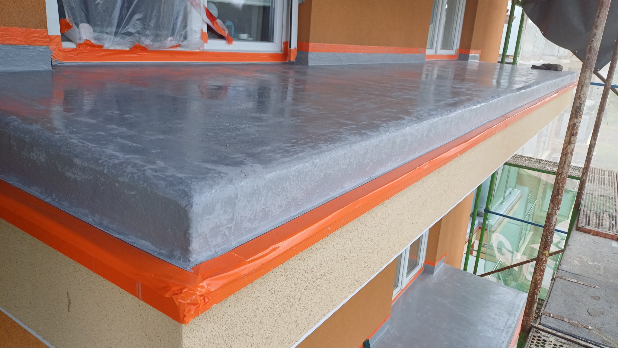 Szara, błyszcząca posadzka na balkonie, zabezpieczona pomarańczową folią malarską, widok z góry. Na drugim planie elewacja budynku w odcieniach beżu i brązu.