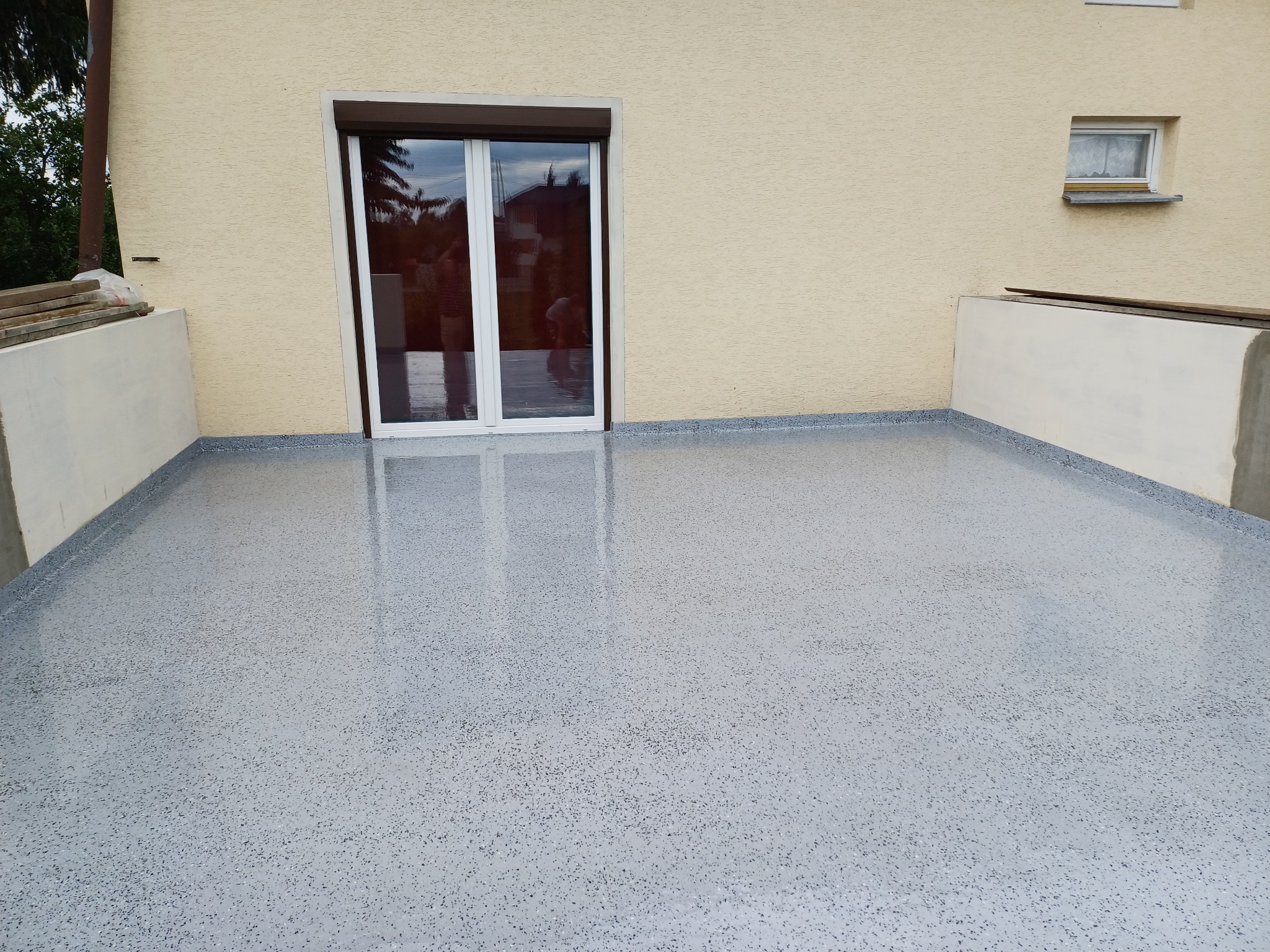 Balkon z szarą, błyszczącą posadzką żywiczną o strukturze lastryko, białe drzwi balkonowe z brązową roletą, kremowa elewacja budynku z małym oknem.