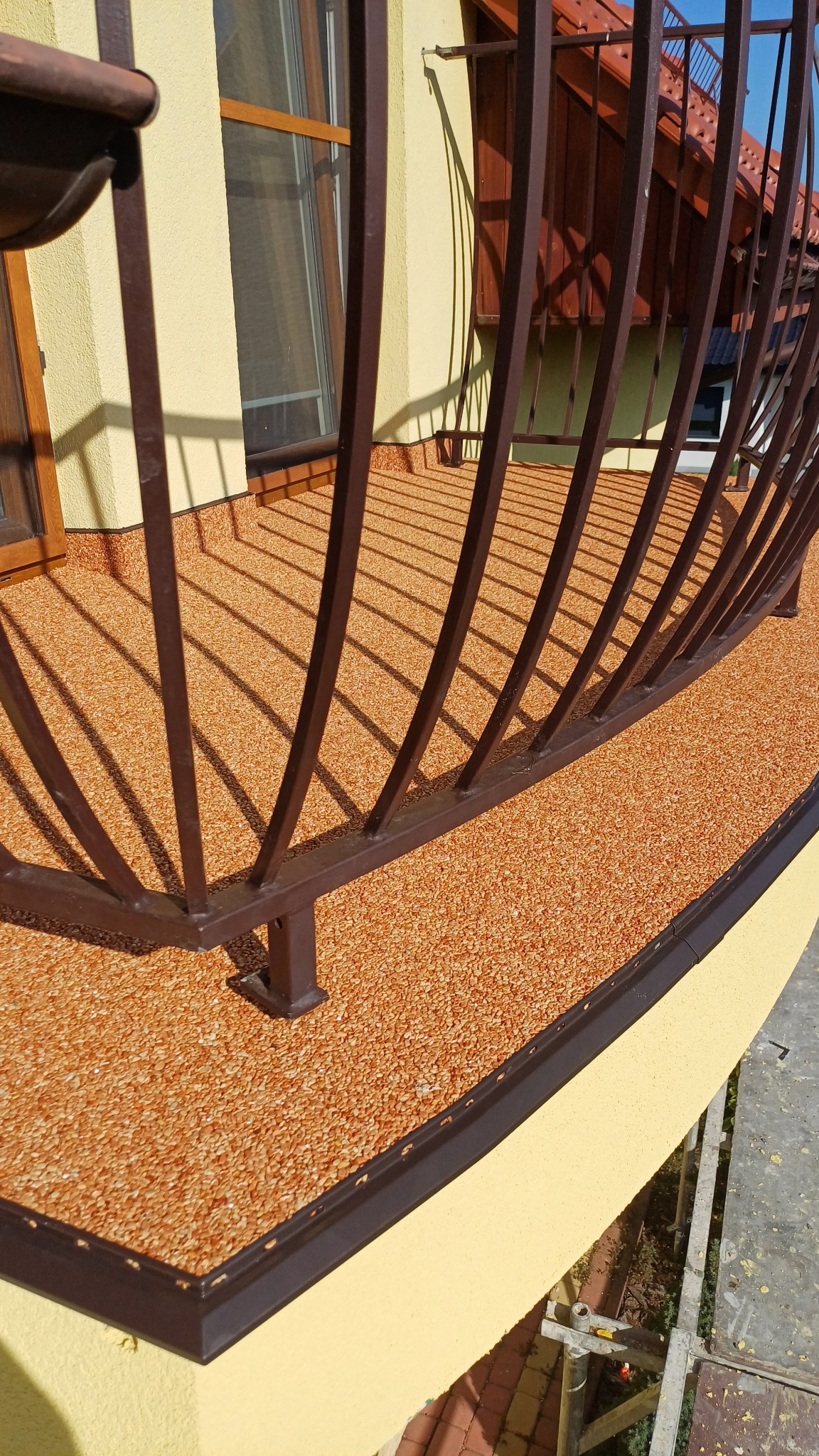 Balkon z brązową balustradą i pomarańczową, żywiczną nawierzchnią. Widoczny fragment elewacji w kolorze kremowym i cień rzucany przez balustradę.