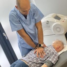 KLINIKA HB Warszawa - Fizjoterapia bólu miednicy - Uroginekologia kobiet i mężczyzn - Fizjoterapeuta w błękitnym uniformie wykonuje masaż pleców starszemu mężczyźnie leżącemu na kozetce. W tle widoczny sprzęt medyczny.