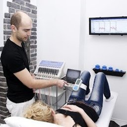 KLINIKA HB Warszawa - Fizjoterapia bólu miednicy - Uroginekologia kobiet i mężczyzn - Mężczyzna w białym fartuchu obsługuje urządzenie do diagnostyki pacjentki leżącej na kozetce. Widoczny monitor z wynikami pomiarów i elementy wyposażenia gabinetu.