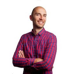 KLINIKA HB Warszawa - Fizjoterapia bólu miednicy - Uroginekologia kobiet i mężczyzn - Uśmiechnięty, łysy mężczyzna w czerwono-niebieskiej koszuli w kratę, z założonymi rękami na białym tle. Portret.