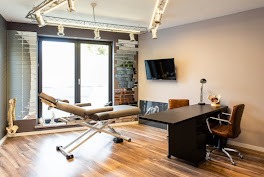 Nowoczesny gabinet rehabilitacyjny: regulowany stół do masażu, biurko z krzesłem, dekoracje ścienne i oświetlenie w ciepłych barwach. Przytulne, profesjonalne wnętrze.