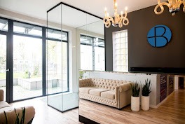 Eleganckie wnętrze recepcji kliniki z beżową sofą chesterfield, szklaną ścianką i widokiem na ogród przez okno. Na ścianie logo kliniki w kolorze niebieskim.