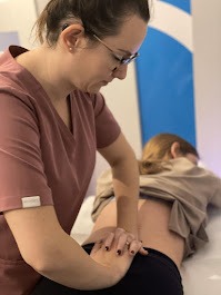 Kobieta w okularach wykonuje masaż dolnej części pleców pacjentki leżącej na stole do masażu w gabinecie. Widoczny niebieski baner w tle.