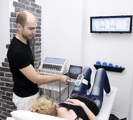 Mężczyzna w białym fartuchu obsługuje urządzenie do diagnostyki pacjentki leżącej na kozetce. Widoczny monitor z wynikami pomiarów i elementy wyposażenia gabinetu.