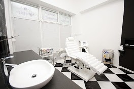 Jasne wnętrze gabinetu z białym fotelem do zabiegów, umywalką i sprzętem medycznym. Czarno-biała szachownica na podłodze. Minimalistyczny, sterylny wystrój.